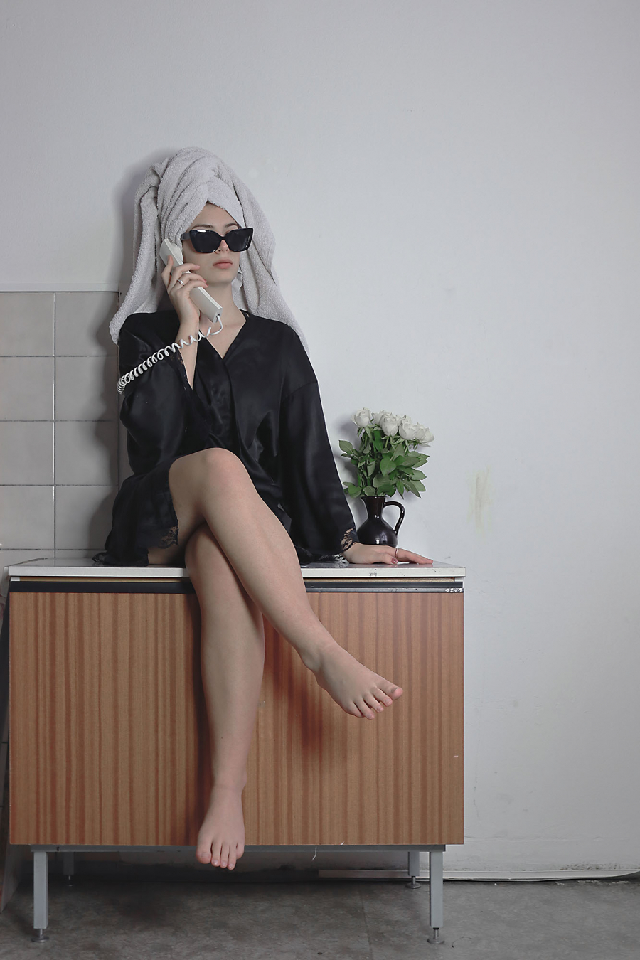 Girl with a towel around her head and black glasses sitting on a cupboard and talking on the phone