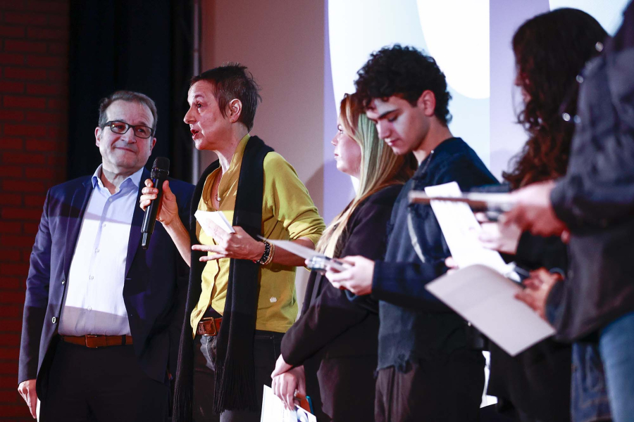 Woman speaking to an audience, man watching, people with certificates on stage